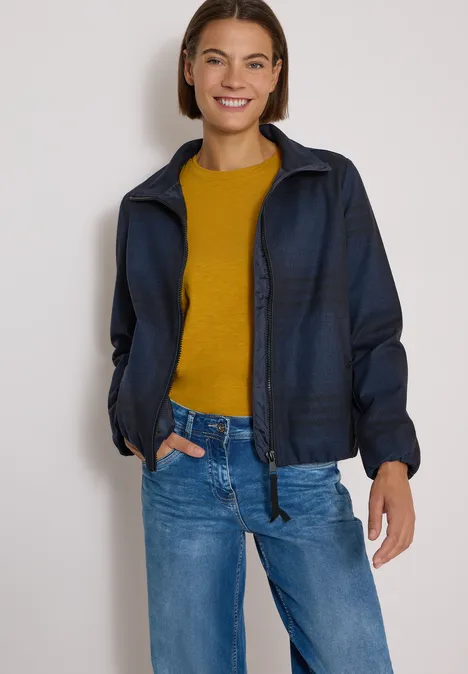 Eine lächelnde Frau trägt eine dunkelblaue Jacke, gelbes Shirt und blaue Jeans.