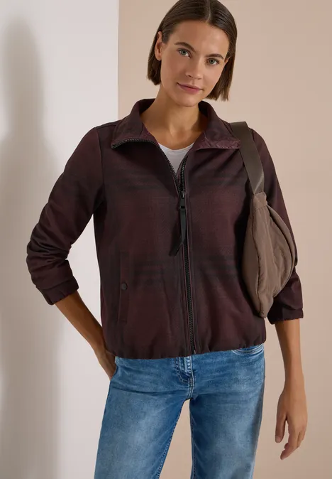 Frau in legerer Jacke, Jeans und Schultertasche lehnt entspannt an einer weißen Wand.