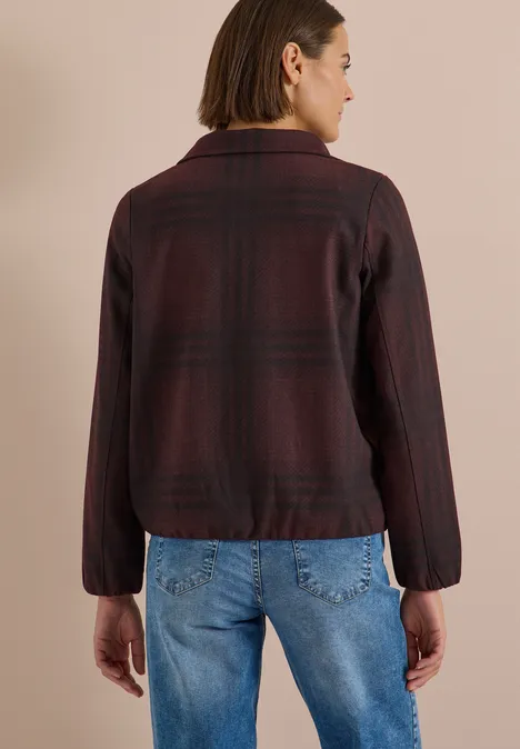 Rückansicht einer Frau mit schulterlangem Haar, die eine karierte, burgunderfarbene Jacke und blaue Jeans trägt.