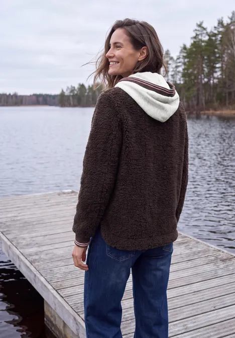 Frau in braunem Fleece-Hoodie steht lächelnd auf einem Steg am See, umgeben von Bäumen und Wasser.