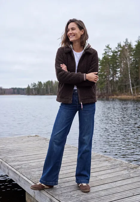 Eine lächelnde Frau mit brauner Jacke, blauen Jeans und Hausschuhen steht mit verschränkten Armen auf einem Steg am See.