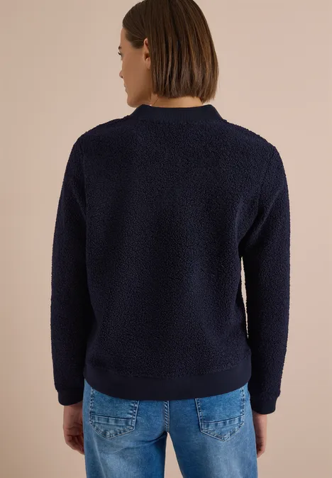 Eine Person von hinten trägt einen dunkelblauen Teddy-Pullover vor beigem Hintergrund.