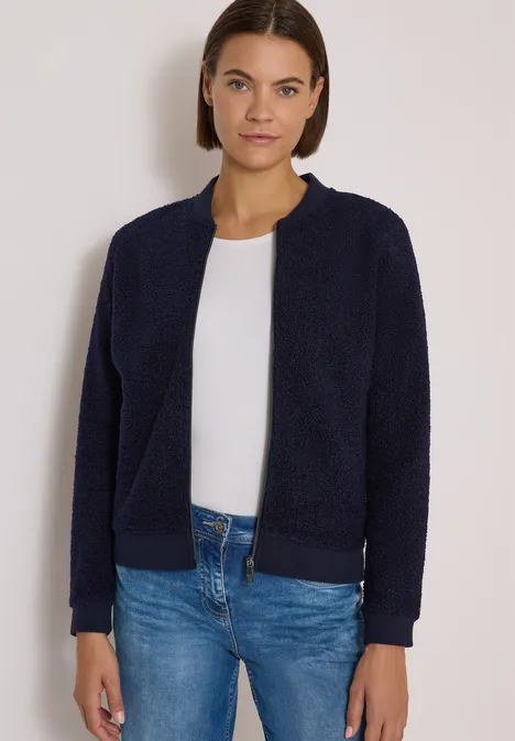 Frau mit kurzen braunen Haaren trägt dunkelblaue Fleece-Jacke, weißes Shirt und blaue Jeans.