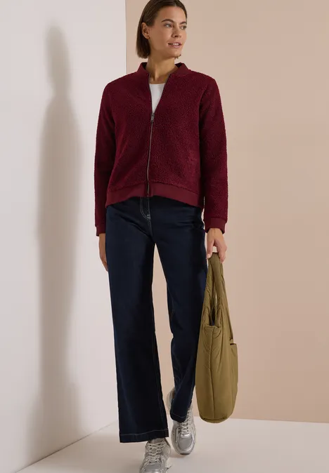 Frau in bordeauxroter Fleecejacke, weiten Jeans und silbernen Sneakers, trägt eine olivgrüne Tasche.