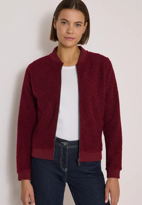 Frau trägt rote Teddyjacke, weißes Shirt und dunkle Jeans vor hellem Hintergrund.