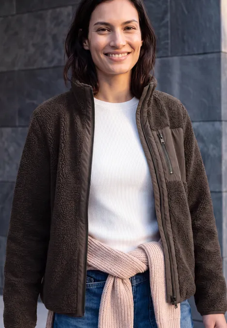 Een glimlachende vrouw draagt een bruine fleecejas, een wit shirt en heeft een vest om haar middel geknoopt.