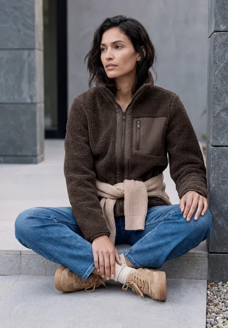 Jonge vrouw zit in kleermakerszit voor een grijs gebouw, draagt een bruine fleecejas, spijkerbroek en beige sneakers.