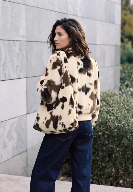 Fake Fur Blouson mit Kuhmuster pearl beige Frau in braun-weißem Fellmantel und blauen Jeans steht seitlich vor einer Steinmauer.