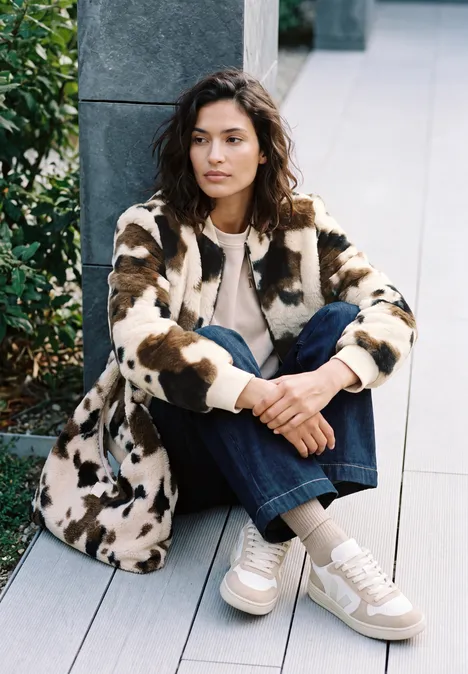 Fake Fur Blouson mit Kuhmuster pearl beige Eine Frau sitzt lässig auf einer Holzterrasse in gemustertem Mantel, Jeans und Sneakers.