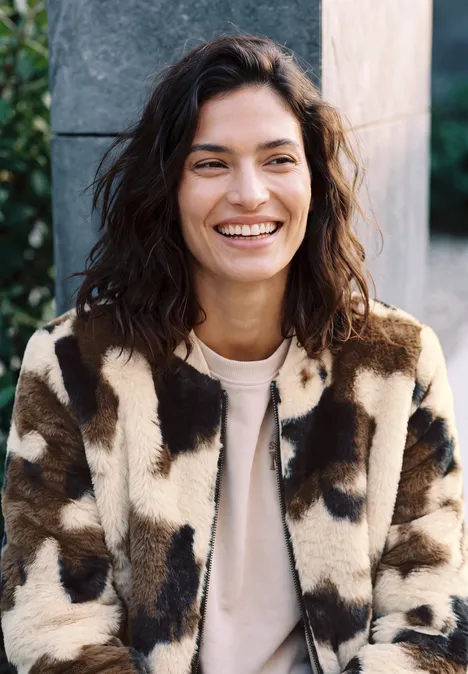 Fake Fur Blouson mit Kuhmuster pearl beige Eine lächelnde Frau in einer braun-weißen, fleckenbemusterten Jacke vor grauer Kulisse
