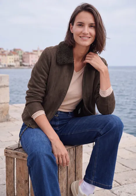 Velours Jacke mit Teddy-Kragen truffle brown Frau in Jeans und brauner Jacke sitzt lächelnd auf einer Kiste am Wasser, im Hintergrund alte Hafenhäuser.