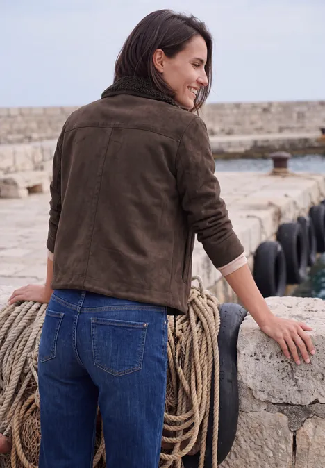 Velours Jacke mit Teddy-Kragen truffle brown Frau in brauner Jacke lehnt am Hafen an einer Steinmauer mit Seil und blickt aufs Meer.