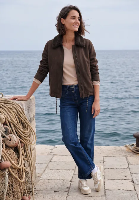 Velours Jacke mit Teddy-Kragen truffle brown Eine lächelnde Frau in Jeans und Jacke lehnt an einer Pierpoller mit Seilen und blickt aufs Meer.