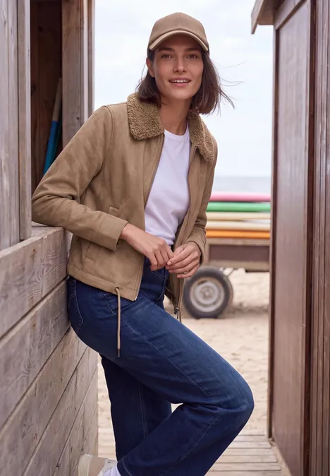 Velours Jacke mit Teddy-Kragen creamy mocha Frau in beiger Jacke, Jeans und Cap lehnt an Holzwand am Strand, im Hintergrund Sand und Meer.
