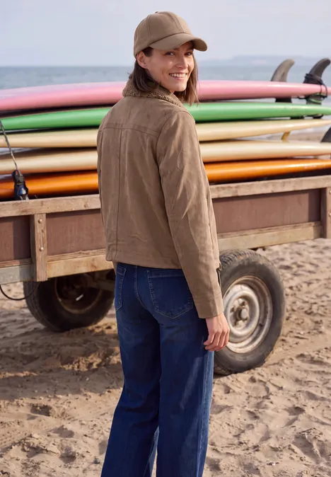 Velours Jacke mit Teddy-Kragen creamy mocha Eine lächelnde Frau in Jacke und Cap steht am Strand neben bunten Surfbrettern.
