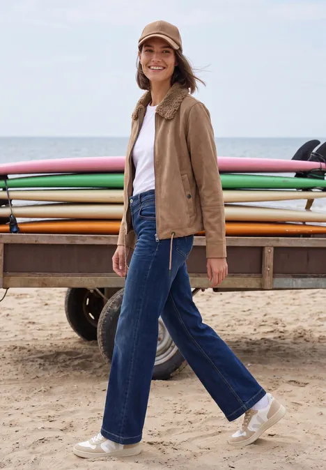 Velours Jacke mit Teddy-Kragen creamy mocha Frau lächelt am Strand vor einem Surfbrettwagen, trägt Jeans, weißes Shirt, braune Jacke und Cap.