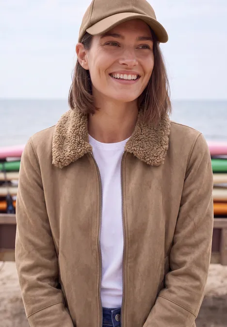 Velours Jacke mit Teddy-Kragen creamy mocha Eine lächelnde Frau am Strand in beigefarbener Jacke mit Teddy-Kragen, weißem T-Shirt und Cap.