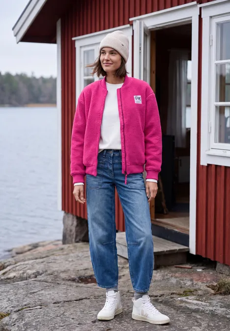 Vrouw in roze jasje, spijkerbroek en muts staat lachend voor een rood huisje aan het meer.