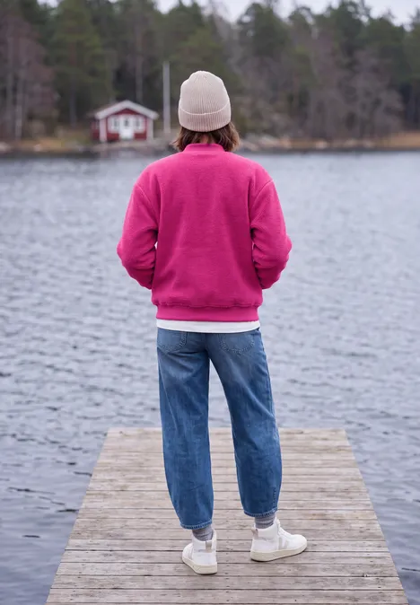 Persoon in roze trui en witte muts staat op steiger, kijkt uit over rustig meer met huisje in bos.