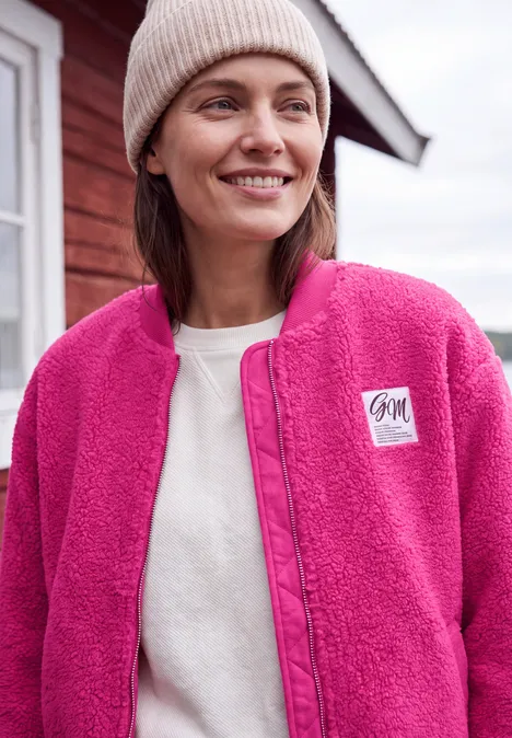 Vrouw lacht in roze fleecejack en muts voor een rood huis.