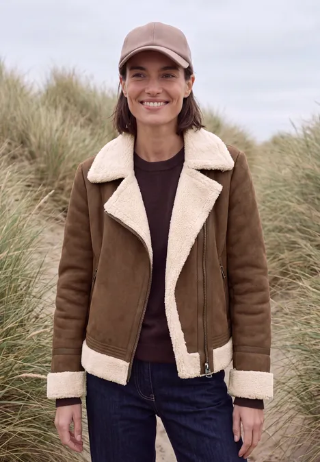 Eine Frau mit brauner Lammfelljacke und Cap steht lächelnd vor grasbewachsenen Dünen.
