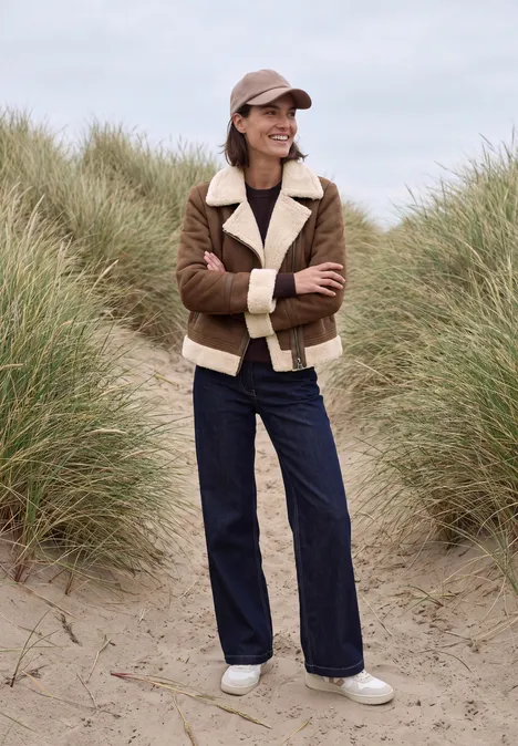 Frau in brauner Lammfelljacke, dunkler Hose und Mütze steht am Strand vor Dünengras.