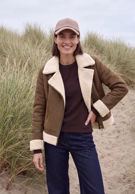 Lächelnde Frau vor Dünen, in Wolljacke, Jeans und Cap.