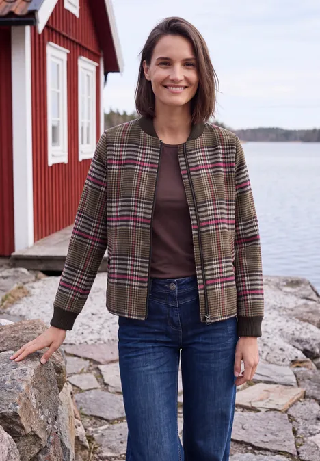 Lächelnde Frau in karierter Jacke und Jeans steht vor rotem Haus am Wasser.
