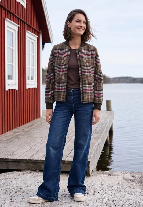 Frau in Karojacke und Jeans steht auf einem Steg neben einem roten Haus am Wasser und lächelt.