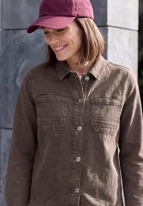 Denim Overshirt mit Brusttaschen truffle brown Lächelnde Frau mit roter Baseballkappe und braunem Hemd mit Brusttaschen vor grauer Mauer.