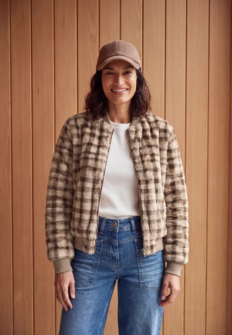 Eine Frau in einer beige-karierten Jacke, weißem T-Shirt, blauen Jeans und Beanie lächelt vor Holzpaneelen.