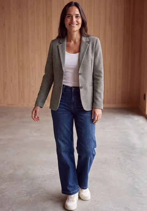 Frau in grauem Blazer, weißem Top, blauen Jeans und weißen Sneakers steht in modernem Raum mit Holzverkleidung und lächelt.
