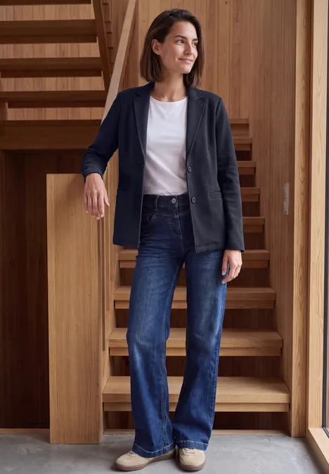 Eine Frau in Jeans, weißem Shirt und dunkelblauem Blazer lehnt lächelnd an einer Holztreppe.
