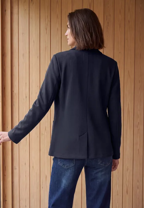 Eine Frau von hinten in schwarzem Blazer und blauen Jeans vor einer Holzlattenwand.