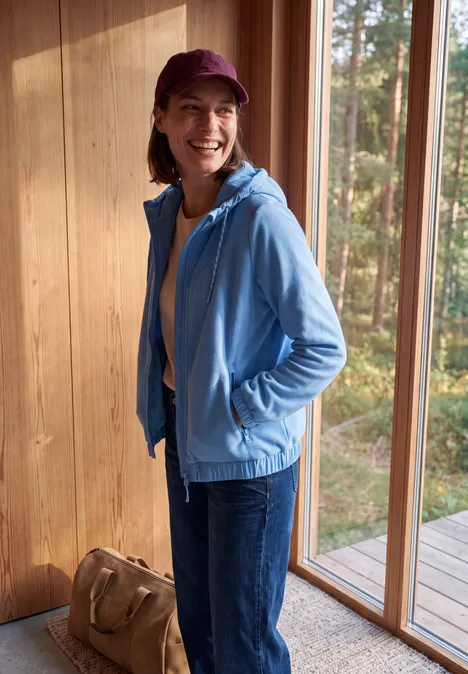 Glimlachende vrouw in blauw vest en jeans met een bordeaux pet, staand bij houten raam met boszicht.