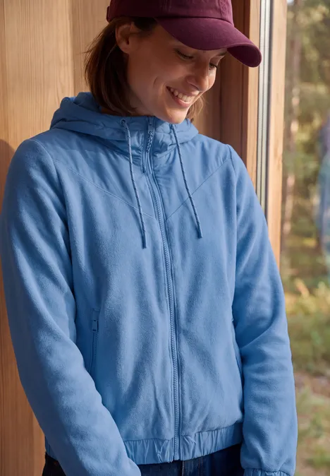 Jonge vrouw in een blauw capuchonvest en pet die glimlacht bij een raam met boszicht.