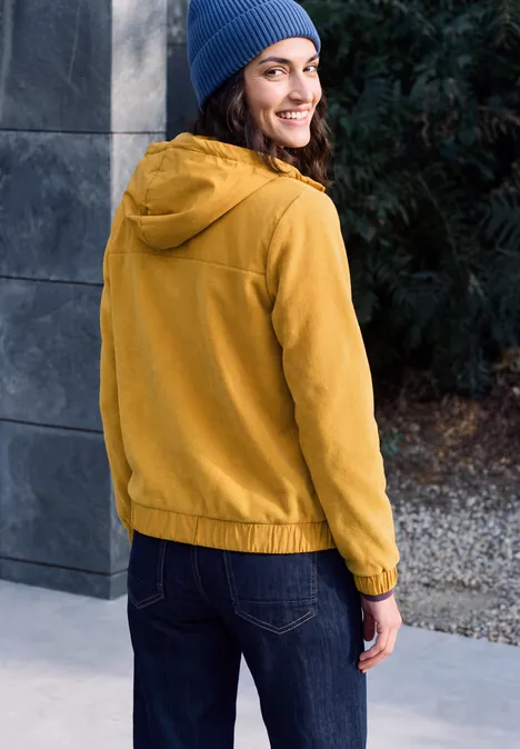 Fleecejas met capuchon en softshell-details golden honey Een vrouw staat buiten in gele jas en blauwe muts, kijkt glimlachend achterom.