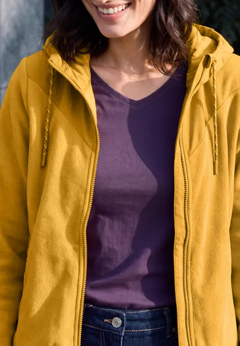 Fleecejas met capuchon en softshell-details golden honey Vrouw met gele capuchonjas over paars T-shirt, alleen bovenlichaam en lichte glimlach zichtbaar.