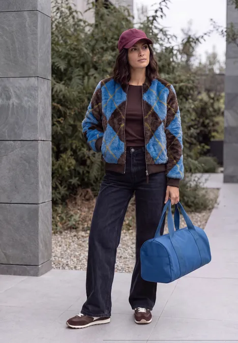 Fake Fur Blouson mit Argylemuster north blue Junge Frau im karierten Mantel, Jeans, Hut und blaue Tasche steht draußen vor einem modernen Gebäude.
