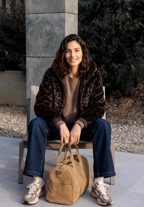 Femme souriante en manteau d'hiver assise sur un muret, tenant un sac de voyage camel.