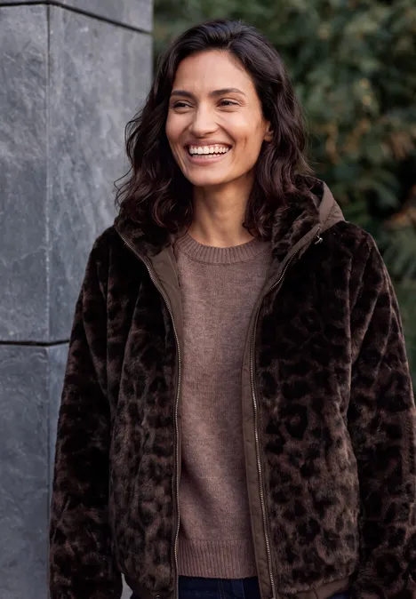 Femme souriante en pull marron et veste pelucheuse foncée devant un mur de pierre et des plantes.