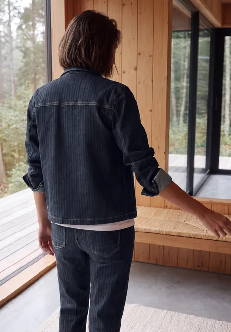 Person von hinten in Jeansjacke und Jeanshose steht in modernem Holzhaus mit großen Fensterfronten.