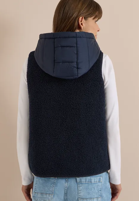 Rückansicht einer Person in weißem Langarmshirt und dunkelblauer, ärmelloser Fleece-Weste mit Kapuze, blaue Jeans.