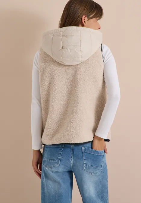 Teddy-Weste mit abnehmbarer Kapuze teddy vanilla white Rückenansicht einer Frau in cremefarbener, wattierter Weste mit Kapuze, weißem Shirt und blauen Jeans.