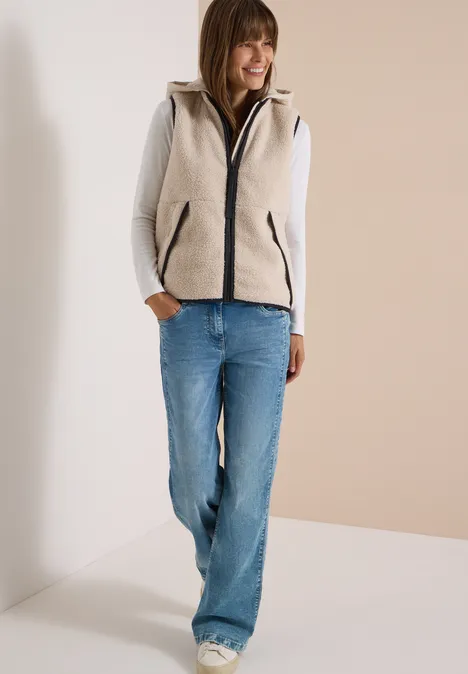 Teddy-Weste mit abnehmbarer Kapuze teddy vanilla white Frau in grauer Fleeceweste mit Kapuze, grauem Langarmshirt und blauen Jeans steht lächelnd mit Händen in Taschen.