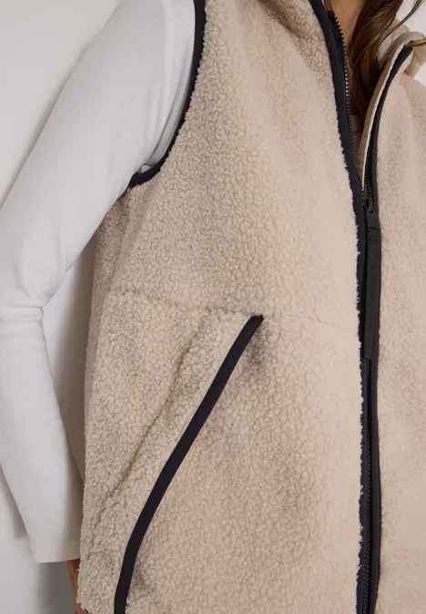 Teddy-Weste mit abnehmbarer Kapuze teddy vanilla white Nahaufnahme einer Person mit weißem Langarmshirt und beiger Fleece-Weste mit schwarzem Reißverschluss.