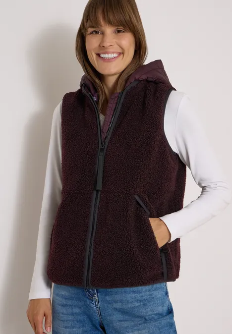 Teddy-Weste mit abnehmbarer Kapuze berry red Eine lächelnde Frau trägt ein weißes Langarmshirt, eine dunkelrote Fleece-Weste mit Kapuze und Jeans.