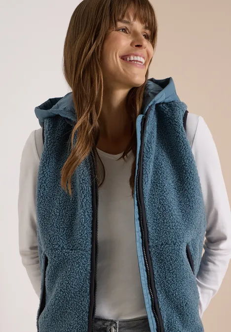 Teddy-Weste mit abnehmbarer Kapuze mountain air blue Frau in weißem Langarmshirt und blauer, ärmelloser Fleece-Weste mit Kapuze, sie lächelt und schaut zur Seite.
