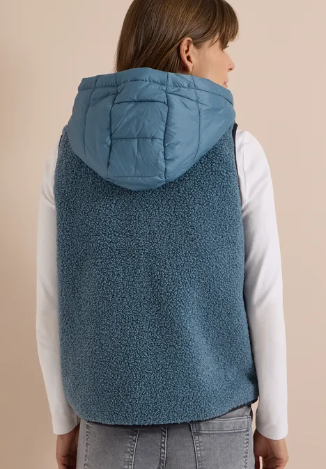 Teddy-Weste mit abnehmbarer Kapuze mountain air blue Frau von hinten in jeansblauer Teddyweste mit gesteppter Kapuze und weißem Langarmshirt.