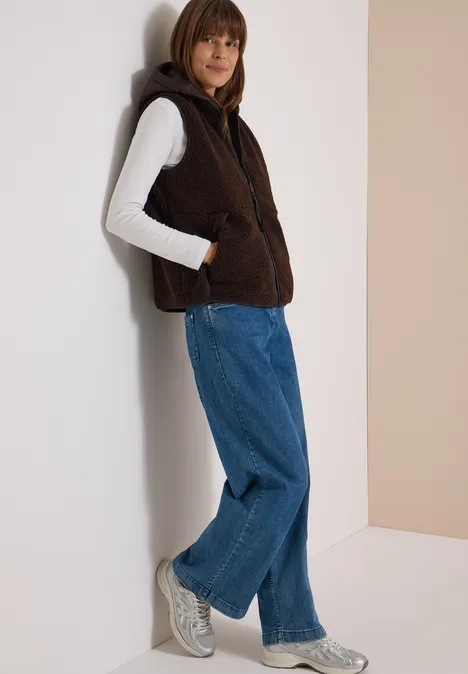 Teddy-Weste mit abnehmbarer Kapuze chocolate sauce brown Frau lehnt an Wand, trägt braune Fleece-Weste mit Kapuze, weißen Pullover, weite blaue Jeans und graue Sneaker.